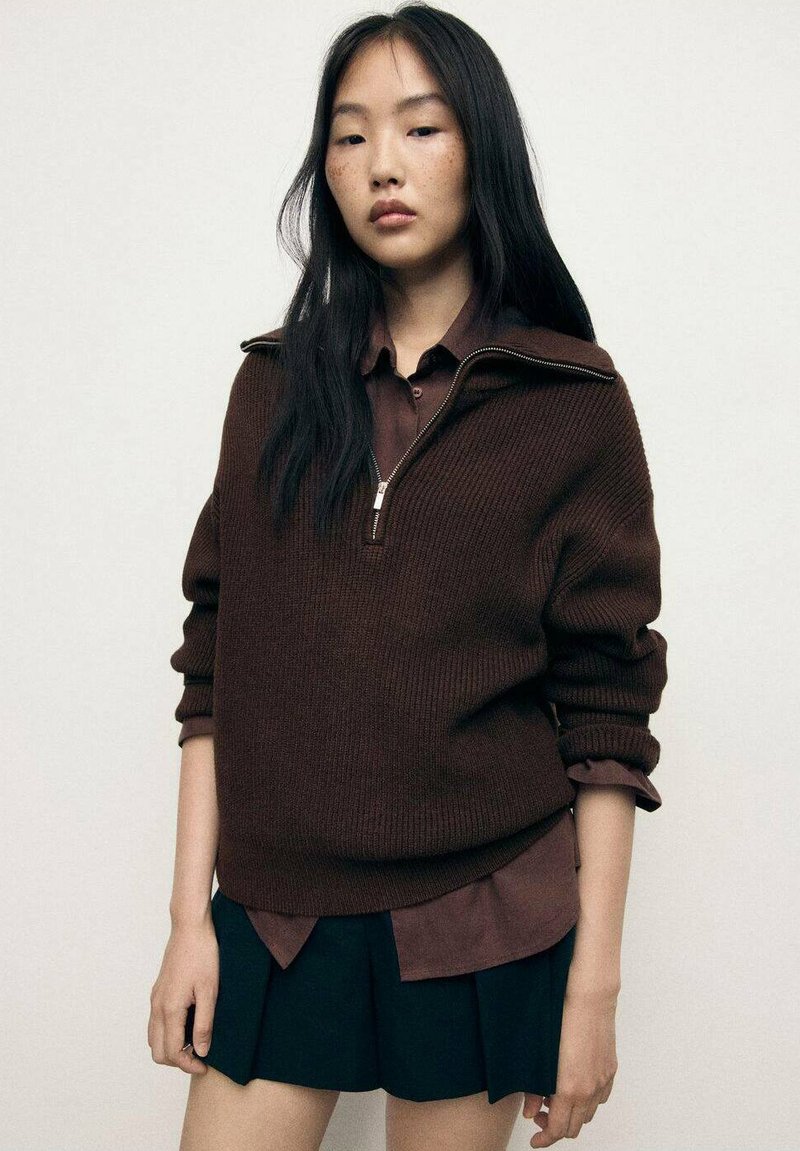 Maglione a coste marrone con collare a mezza zip, indossato sopra una camicia con colletto marrone scuro, abbinato a una gonna nera plissettata.