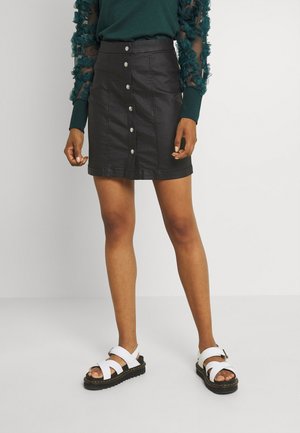 Jupe en denim noir en forme de A avec des boutons froncés et une texture lisse. Associée à des sandales blanches à lanières avec une semelle épaisse.