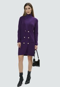 Cardigan long en tricot violet avec col roulé, devant à double boutonnage, deux poches et accents de boutons dorés, porté avec des bottines noires.