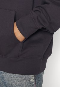 Mano en el bolsillo de una sudadera gris oscuro, combinada con jeans de mezclilla azul claro desgastados, mostrando la textura de la tela y los detalles de la costura.