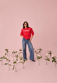 Femme portant un pull rouge "Amour" et un jean, debout parmi des roses roses dans des vases en verre sur un sol et un arrière-plan roses assortis.