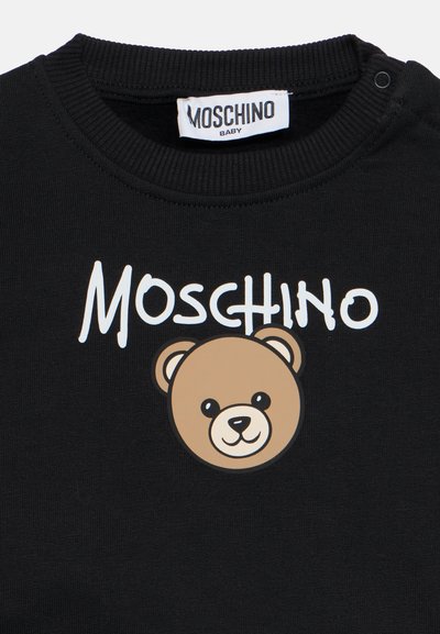 Gros plan sur un t-shirt bébé noir avec le texte "Moschino" et un visage d'ours en peluche souriant imprimé sur le devant sous un col côté.