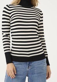 Femme portant un pull à manches longues rayé horizontalement noir et blanc et un jean en denim bleu, debout avec une main relâchée le long du corps.