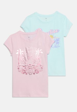 Deux t-shirts enfants à manches courtes, l’un rose avec un motif brillant de soleil et de palmier, l’autre bleu clair avec une impression pastel « Paradise » et palmiers.
