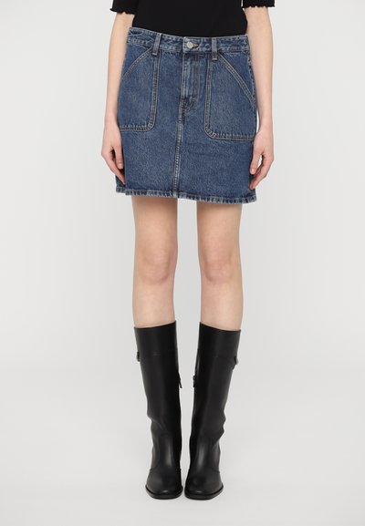 Jupe en denim bleu foncé, avec une taille haute, deux poches avant et un ourlet effiloché. Assortie à des bottes en cuir noir hauteur genou.
