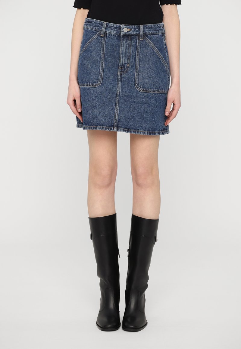 Saia de ganga em azul escuro, com uma cintura alta, dois bolsos frontais e uma bainha desfiada. Combinada com botas pretas de couro até ao joelho.