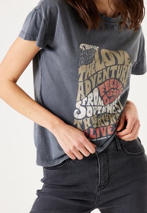 Osoba nosząca wyblakły ciemnoszary T-shirt z kolorowym, wyraźnym tekstem w stylu vintage oraz ciemne spodnie, ręce poprawiające brzeg koszulki.