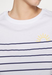 White t-shirt with navy horizontal stripes, featuring a small yellow sun graphic on the left chest. Soft cotton material and crew neckline.