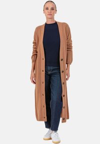 Lang geschnittener Strickcardigan in Kamelhaarfarbe mit geripptem Kragen, Knopfverschluss und langen Ärmeln, getragen über einem dunkelblauen Oberteil und Jeans, kombiniert mit weißen Turnschuhen.