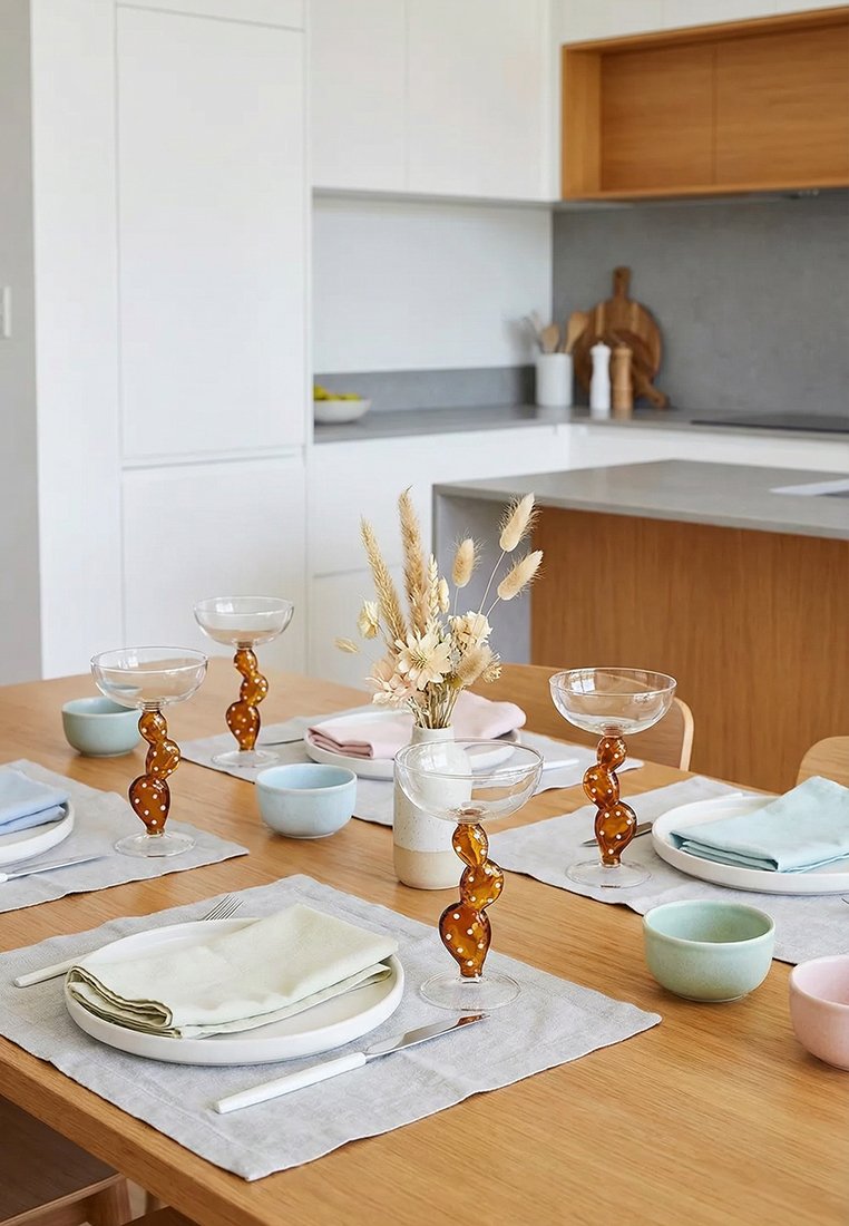 Stół jadalny nakryty dla czterech osób, z kieliszkami o skręconych bursztynowych nóżkach, pastelowymi miseczkami, tekstylnymi serwetkami oraz wazonem z suszonymi kwiatami w nowoczesnej kuchni.