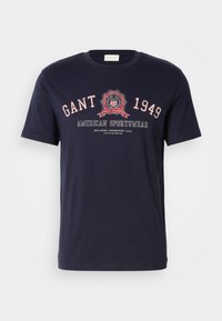 Tricou casual din bumbac bleumarin, cu guler la bază, având text alb și roșu și un logo circular cu "GANT," "1949," și "AMERICAN SPORTSWEAR."