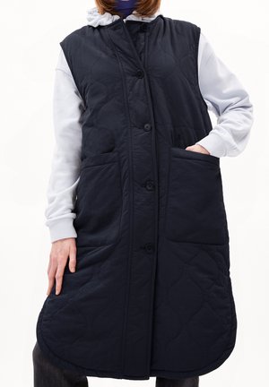 Persoon draagt een lange, zwarte doorgestikte mouwloze bodywarmer met grote voorzakken en knopen, gedragen over een lichtgrijze hoodie.