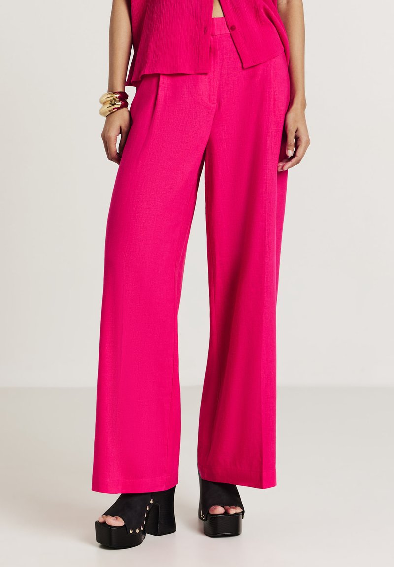 Donna che indossa pantaloni a gamba larga di un vivace rosa, un top abbinato, scarpe con plateau aperte nere e bracciali d'oro su uno sfondo neutro.