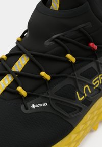 Melns sporta apavu ar Gore-Tex etiķeti, gumijas zoli un dzelteniem akcentiem. Auklu sistēma dažādās krāsās, teksturēts augšdaļas dizains.