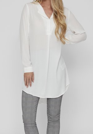 Chemise blanche à manches longues avec col, dotée d'un design unique à ourlet asymétrique et d'un tissu doux et transparent. Assortie à un pantalon à carreaux gris.