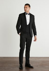 Czarny smoking z satynowymi klapami, z białą koszulą i muszką. Dopasowany krój, eleganckie spodnie i wypolerowane czarne buty. Tło z drewnianej podłogi.