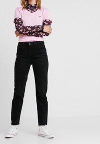 En rosa v-ringad t-shirt över en svart blommig långärmad topp, kombinerad med svarta jeans och vita sneakers med fransad fåll.