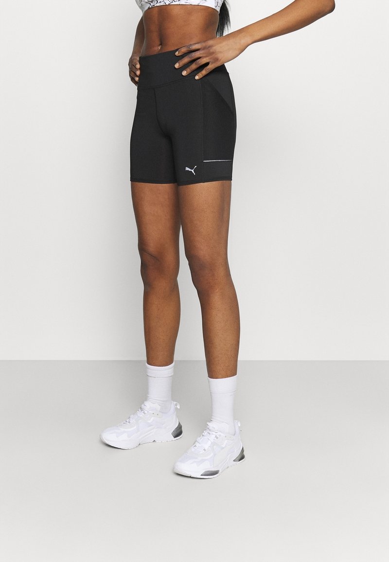 Femme debout, mains sur les hanches, portant un short de sport noir Puma, des baskets blanches et des chaussettes blanches, sur un fond uni.