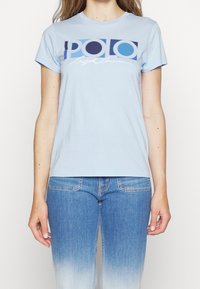 Světle modré bavlněné tričko s krátkým rukávem, které obsahuje výrazné kontrastní logo "POLO" v modré a tmavě modré barvě, spolu se stylizovaným písmem.