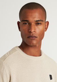 Beiger Pullover mit einem strukturierten Strickmuster, geripptem Ausschnitt und einem kleinen braunen Badge mit einem dreieckigen Design auf der Brust.