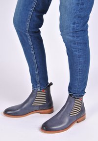 Skórzane botki za kostkę w granatowym kolorze z elastycznymi paskami w paski, z czerwonym serduszkiem. Drewniana pięta, gładka faktura, zestawione z niebieskimi obcisłymi dżinsami.