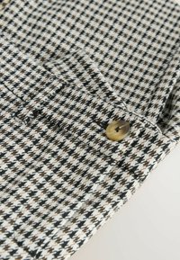 Houndstooth-mønstrede stof i sort, hvid og brun med en glat tekstur. Har en stor, rund, gylden knap på kraven.