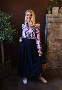 Kwiatowa bluzka z różowo-zielonym wzorem, zestawiona z zwiewną granatową spódnicą. Kobieta stoi w rustykalnym otoczeniu z roślinami i ceglanym murem.