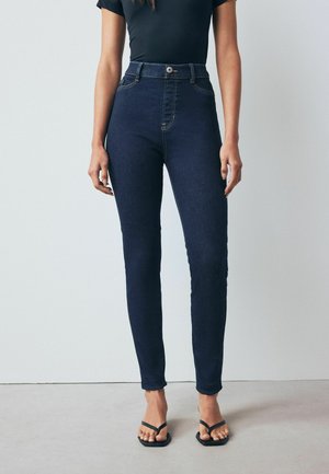 Højtaljede mørkeblå denim jeans med slim fit, der har en frontknap, diskrete stikninger og en glat tekstur. Sammen med sorte sandaler.