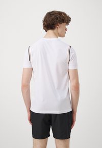 Camiseta atlética blanca de manga corta, con rayas negras y doradas en los hombros; combinada con pantalones cortos negros. Textura de tela suave.