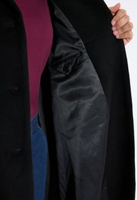 Abrigo de lana negro con un forro de satén suave. El interior cuenta con un bolsillo y logotipos sutiles. Parte superior burgundy visible y jeans azules.