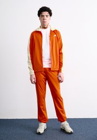 Πορτοκαλί φόρμα με κρέμ λεπτομέρειες; περιλαμβάνει ένα ζακέτα με φερμουάρ και ταιριαστά παντελόνια, κατασκευασμένα από λεία, γυαλιστερή ύλη. Άσπρα αθλητικά υποδήματα ολοκληρώνουν την εμφάνιση.
