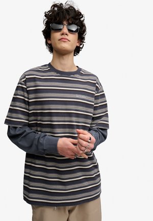 Ung mand med krøllet hår iført solbriller, en stribet kortærmet skjorte over en langærmet top og beige bukser, justerer sine ringe.