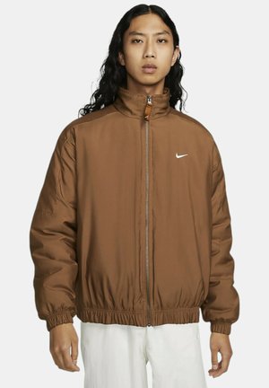 Veste rembourrée marron avec un col haut, fermeture éclair sur le devant, et poignets et ourlet élastiqués. Présente un petit logo Nike blanc sur la poitrine.