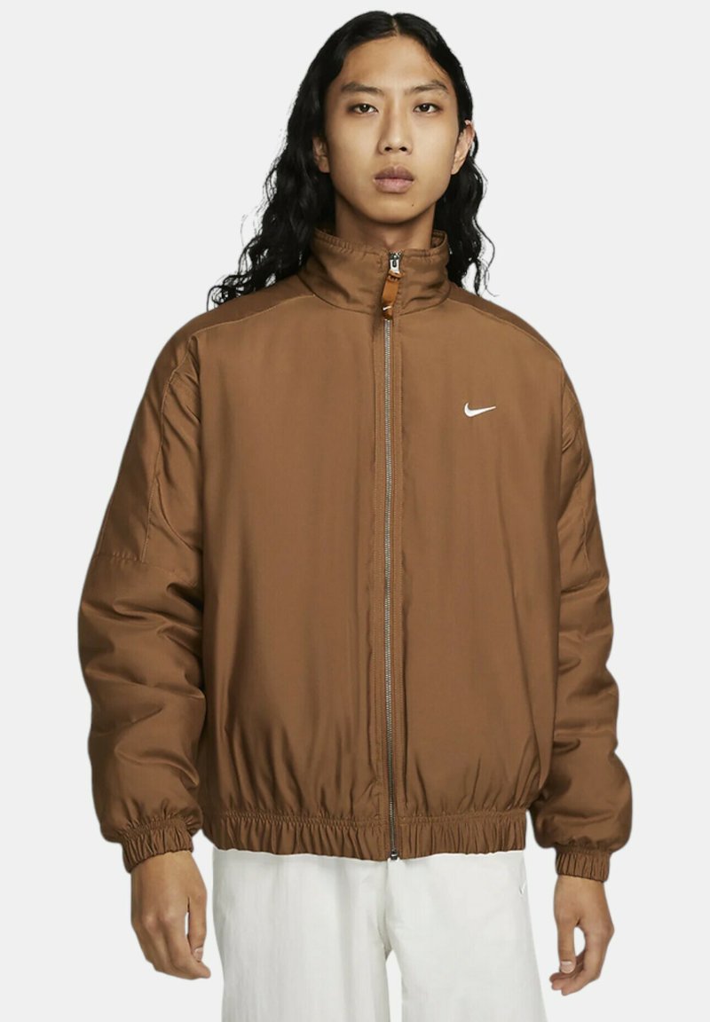 Hnědá prošívaná bunda s vysokým límcem, předním zipem a elastickými manžetami a spodním lemem. Obsahuje malé bílé logo Nike na hrudi.