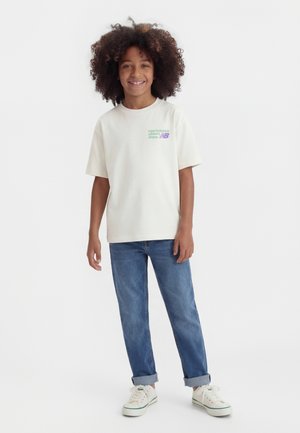 Bambino sorridente con capelli ricci che indossa una maglietta bianca New Balance, jeans blu risvoltati e scarpe da ginnastica bianche, in piedi davanti a uno sfondo semplice.