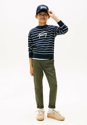 Marine gestreepte trui met witte lijnen en logo, olijfgroene broek en beige sneakers. De jongen draagt een bijpassende marineblauwe pet en lacht.