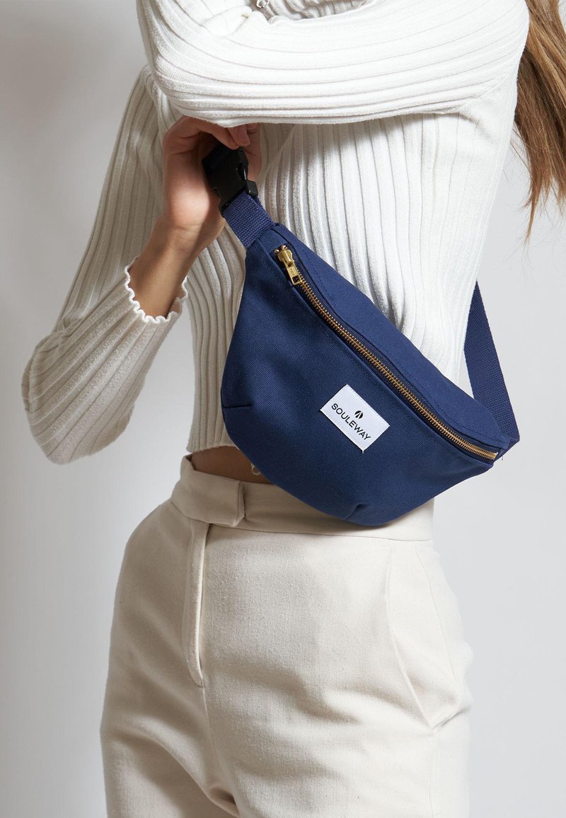 Riñonera de tela azul con una cremallera dorada, que presenta una etiqueta rectangular blanca en la parte frontal. Llevada al hombro.