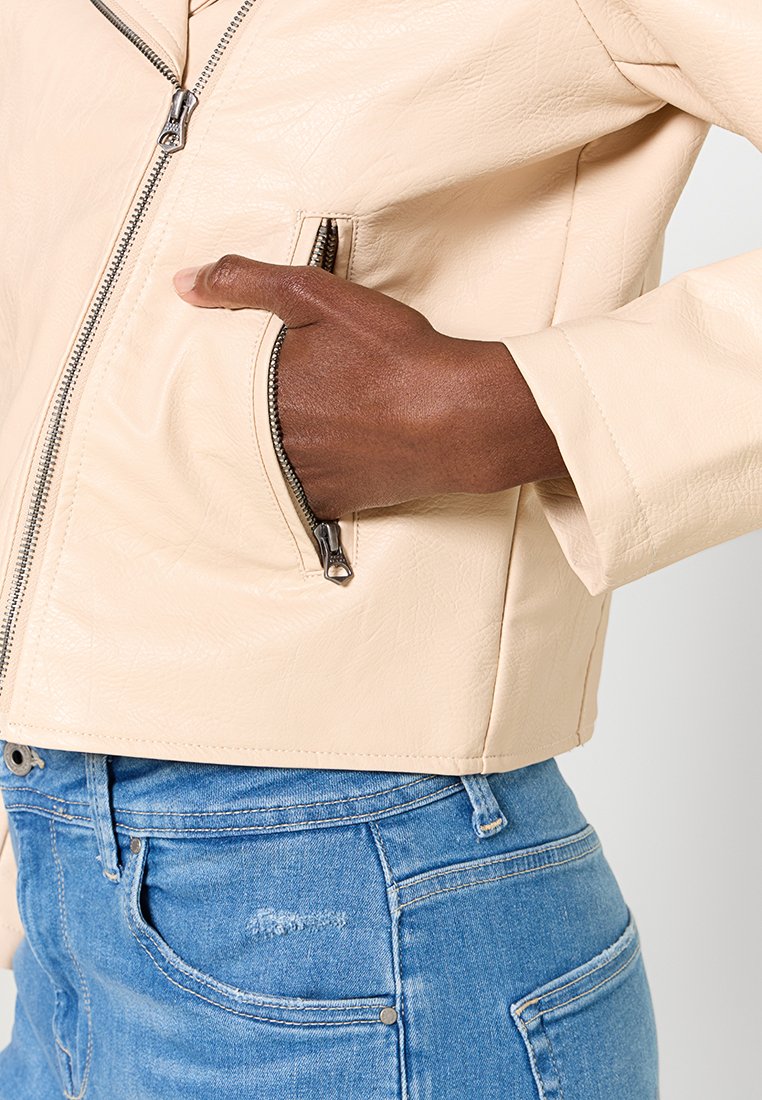Chaqueta de piel sintética color beige, diseño corto, detalle de cremallera visible, bolsillo lateral, textura suave, combinada con jeans de denim azul.