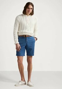 Νέος άνθρωπος που στέκεται φορώντας ένα λευκό πουλόβερ με πλεξούδα, μπλε σορτς με καφέ ζώνη και λευκά αθλητικά παπούτσια σε έναν απλό φόντο.