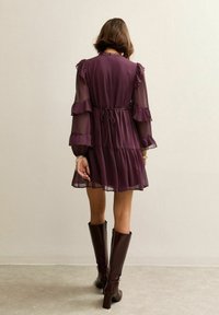 Vrouw die een doorschijnende, lange mouwen paarse jurk met ruches draagt, gecombineerd met donkerbruine laarzen tot net onder de knie, staand tegen een effen lichte muur.