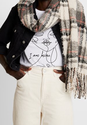 Personne portant un pantalon blanc, une veste en jean noire, un t-shirt blanc avec un visage abstrait et le texte « Je suis à moi », ainsi qu'une écharpe à carreaux.