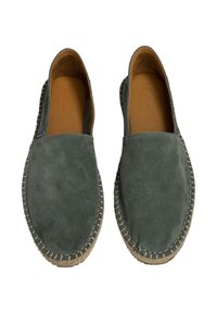Scarpe slip-on in suede verde con punta rotonda e suola in juta intrecciata. L'interno presenta un rivestimento liscio color cammello. Design minimalista senza hardware visibile.