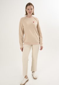 Felpa beige con un piccolo cuore rosso sul petto, abbinata a pantaloni dritti off-white e sneakers bianche con dettagli beige.