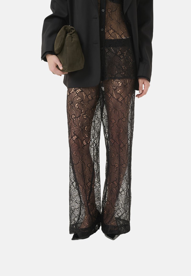 Schwarze Spitzenhose mit floralem Muster, mit einem transparenten Design und weitem Bein. Begleitet von einem schwarzen Blazer und einer olivgrünen Tasche.