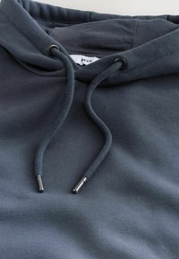 Pilkas džemperis su švelniu audiniu, turintis kapišoną su raišteliais ir metaliniais antgaliais. Paprasto dizaino, be matomų raštų ar logotipų.