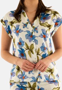 Camisa de botones con estampado floral que presenta flores azules y naranjas sobre un fondo blanco, con mangas cortas y un corte relajado. Textura suave.