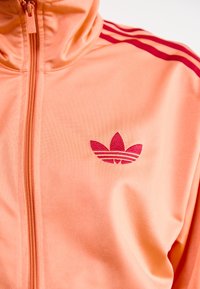 Helles orange Zip-Up-Jackett mit roten drei-Streifen-Akzenten. Verfügt über einen strukturierten Stoff und ein kleines pinkes Adidas-Trefoil-Logo auf der Brust.