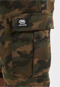Sot shorts cargo kamuflase dalam warna coklat dan hijau, dilengkapi dengan saku tempel yang memiliki label logo hitam. Terbuat dari kain bertekstur dengan potongan yang santai.