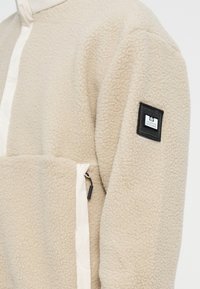 Beige fleece genser med en glatt, lysfarget panel ved nakken, glidelåslommer og en svart logo-patch på armen.