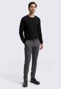 Mann trägt einen schwarzen Langarmpullover, graue Slim-Fit-Hosen mit schwarzem Gürtel und schwarze Lederstiefel, steht vor einem einfarbigen hellen Hintergrund.